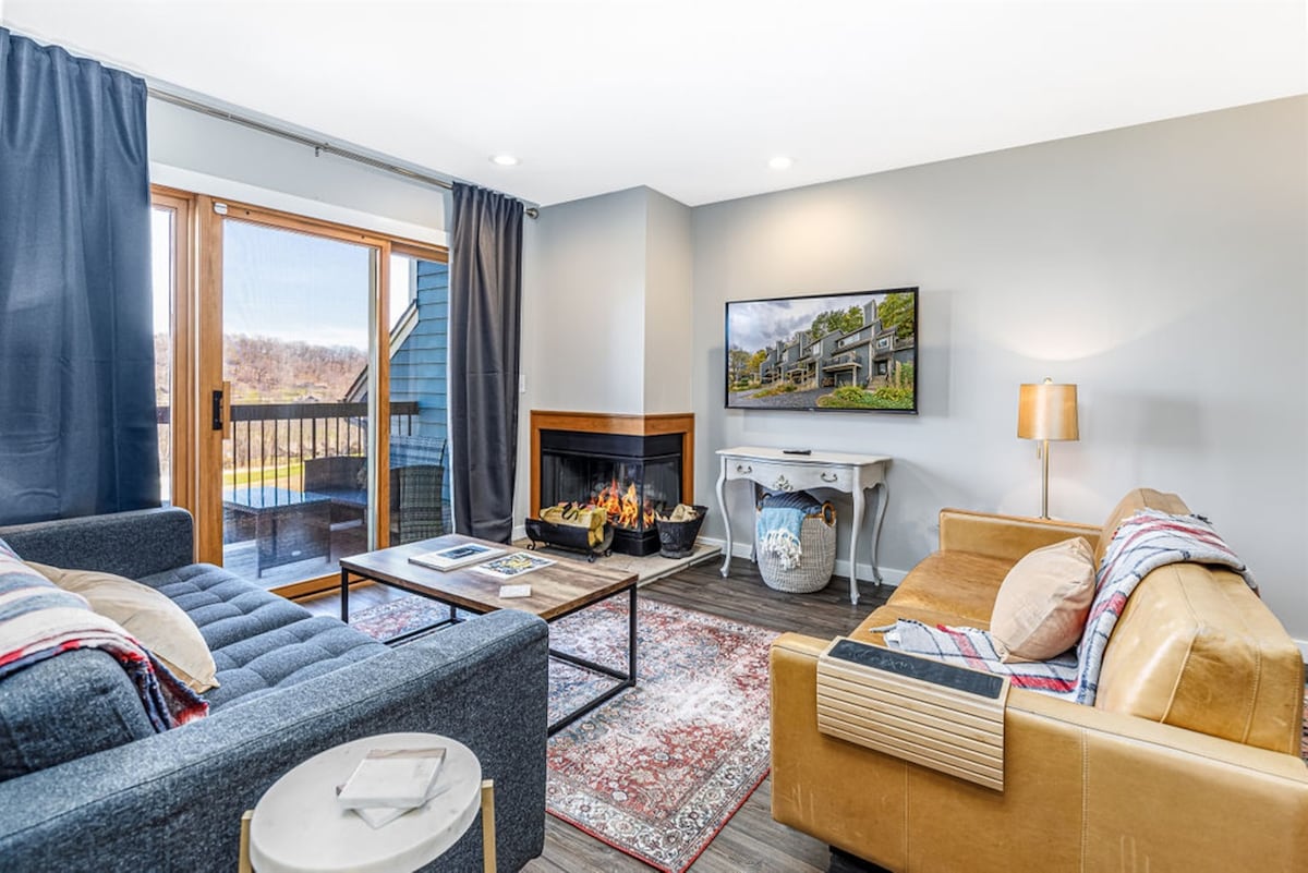 A living area is showcased with modern furnishings, including a dark blue sofa and a tan leather sofa. A wood-burning fireplace is set within a decorative surround, and a large television is mounted on the wall. Natural light enters through sliding glass doors leading to a private patio.