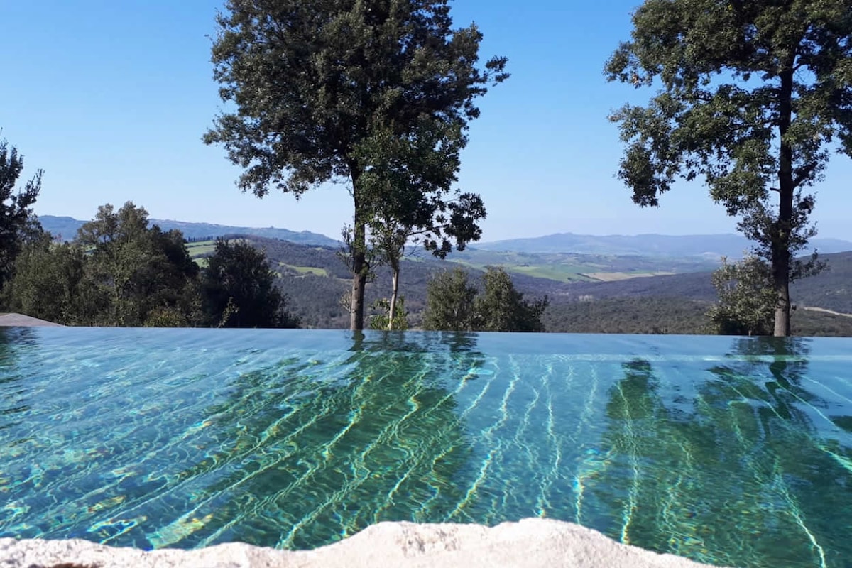 An infinity swimming pool overlooks rolling hills and lush countryside, with clear blue skies above. The serene water reflects the surrounding trees, enhancing the sense of tranquility and connection to nature.