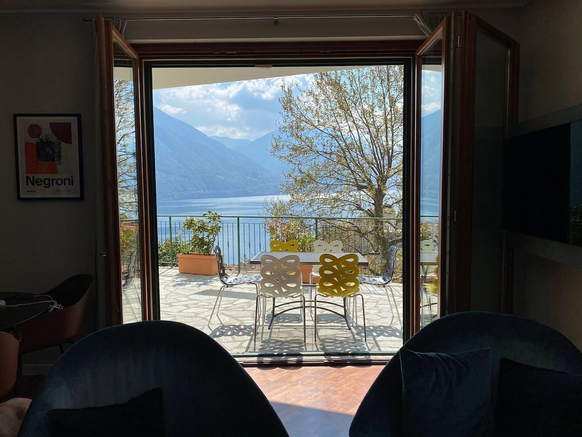 A view from within the living area showcases double glass doors opening to a terrace overlooking the lake. A dining table with colorful chairs is positioned on the stone patio, surrounded by greenery and framed by distant mountains under a partly cloudy sky.