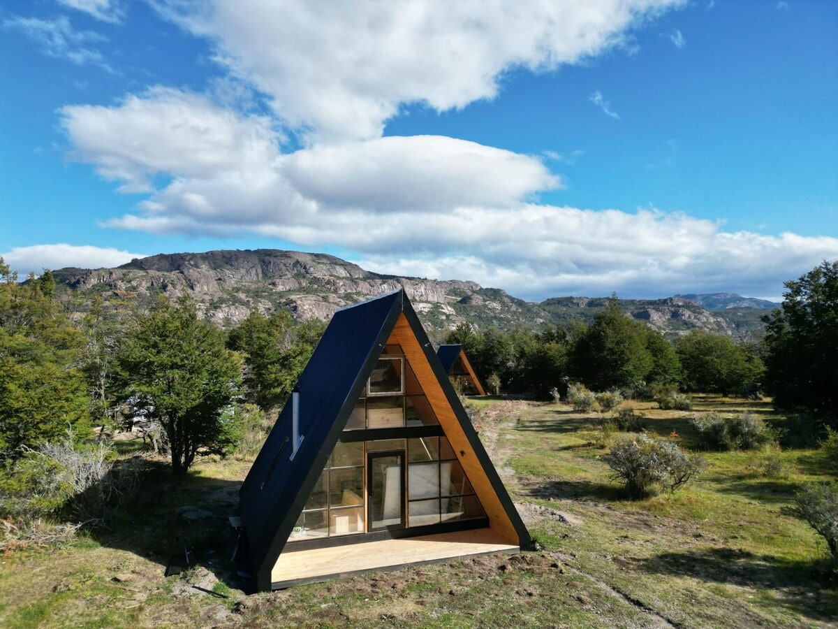 A modern A-frame cabin is set against a backdrop of mountains and blue skies. Large glass windows reflect the surrounding nature, while lush greenery and scattered bushes frame the structure, creating a serene and inviting outdoor setting.