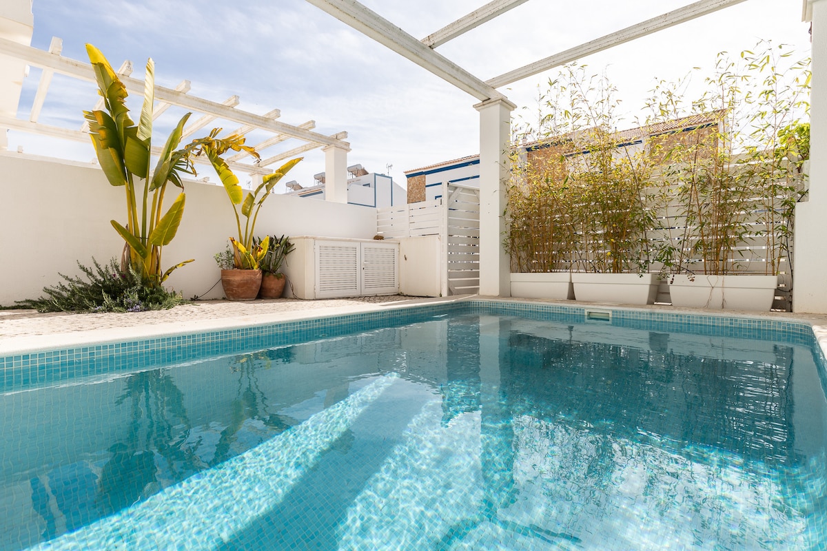 A private swimming pool is surrounded by a patio area featuring greenery, including banana plants and bamboo. The pool's clear water reflects the sky, while the surrounding white walls create a serene atmosphere for relaxation.
