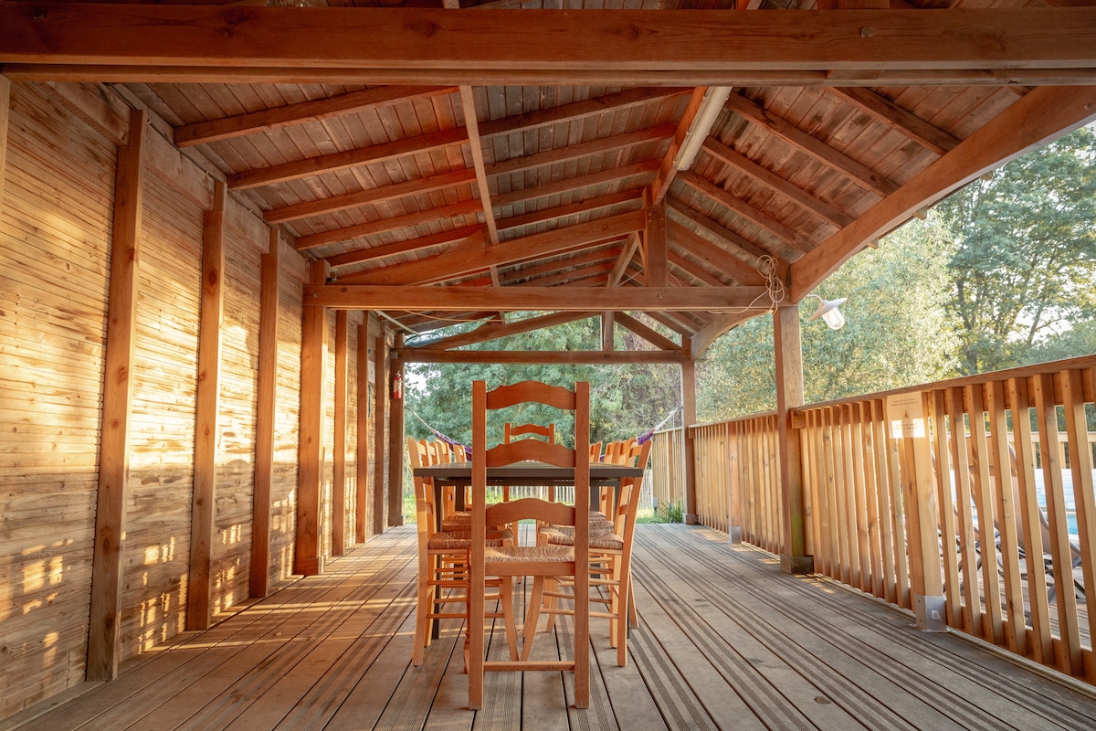 An outdoor terrace is presented under a wooden structure with an arched ceiling. A large dining table with several wooden chairs is centrally positioned. Natural light floods the space, highlighting the warm tones of the wood and the surrounding greenery through the railing.