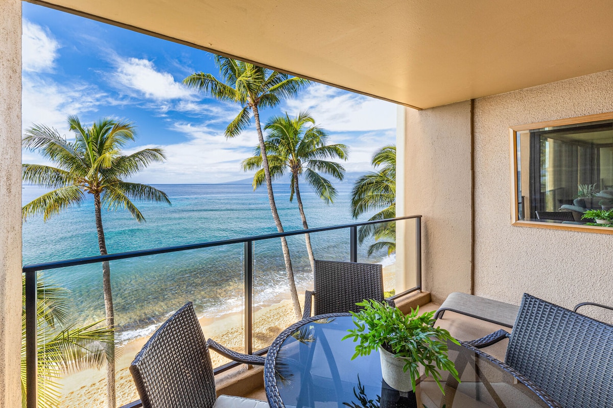 An outdoor lanai features a round glass table surrounded by four woven chairs. Palm trees sway gently in the breeze, framing unobstructed ocean views. The tranquil water reflects a clear blue sky with a few wispy clouds, providing a serene backdrop to the coastal scene.