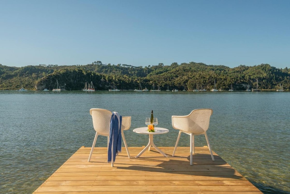 A wooden dock extends over calm waters, featuring two white chairs paired with a small round table. A blue towel sits on one chair, while a bottle of champagne and a glass are placed on the table, with lush green hills and boats visible in the background.