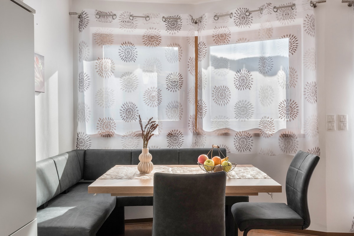 A dining area features a rectangular table surrounded by a mix of cushioned chairs and a comfortable bench. Light filters through sheer curtains adorned with circular patterns, providing a soft ambiance. A decorative vase with dried stems and a bowl of fresh fruit add a touch of warmth.