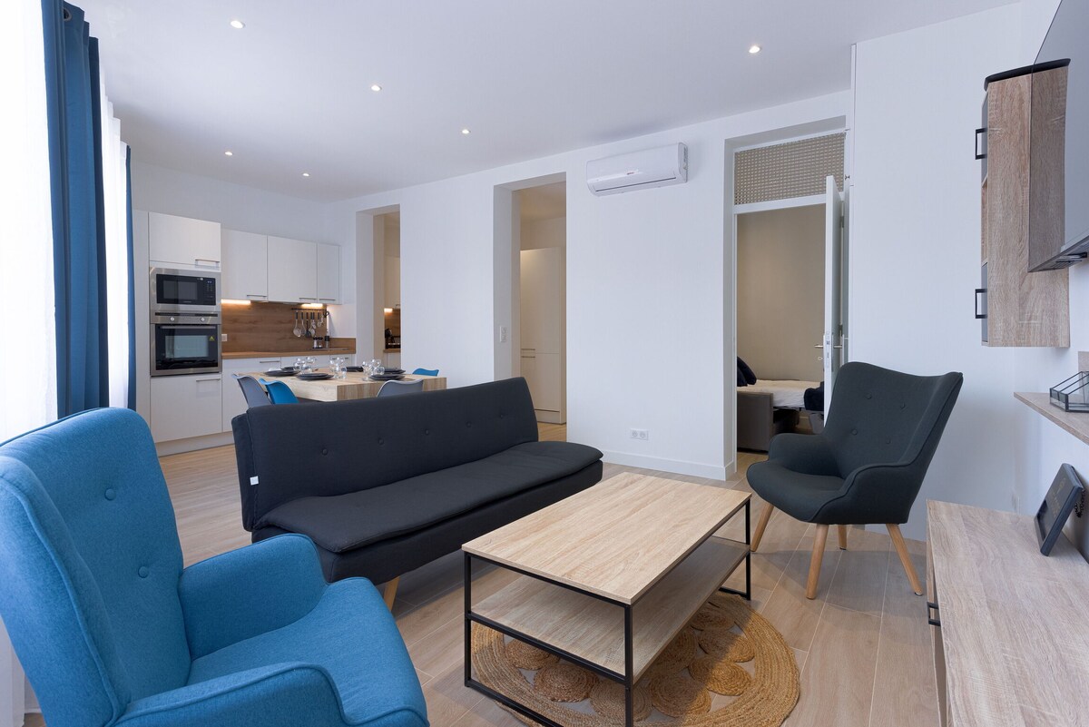 The spacious living area features a modern design with a comfortable black sofa and two blue armchairs. A light wood coffee table sits centrally, complemented by an inviting rug. An open kitchen is visible, showcasing sleek cabinetry and essential appliances, with a dining area situated in the background.