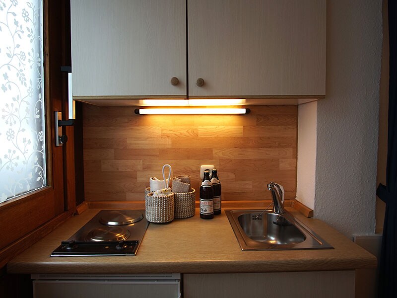 A compact kitchen area features a small stove and a stainless-steel sink under a warm light fixture. Wooden cabinetry provides storage, while a woven basket and bottles are displayed on the countertop, enhancing a functional cooking space.