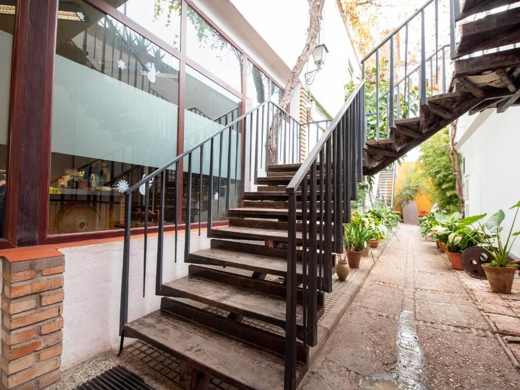An outdoor staircase with a curved black railing leads to an upper level. Surrounding greenery is visible in terracotta pots, while a narrow pathway lined with plants extends alongside a glass-walled area of the building, providing a glimpse of the inviting atmosphere.