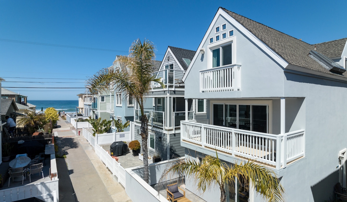 A charming three-story home is pictured with a light-colored exterior and multiple balconies. Palm trees line the walkway leading to the beach. The ocean can be seen in the background under a clear blue sky, providing a serene coastal setting.