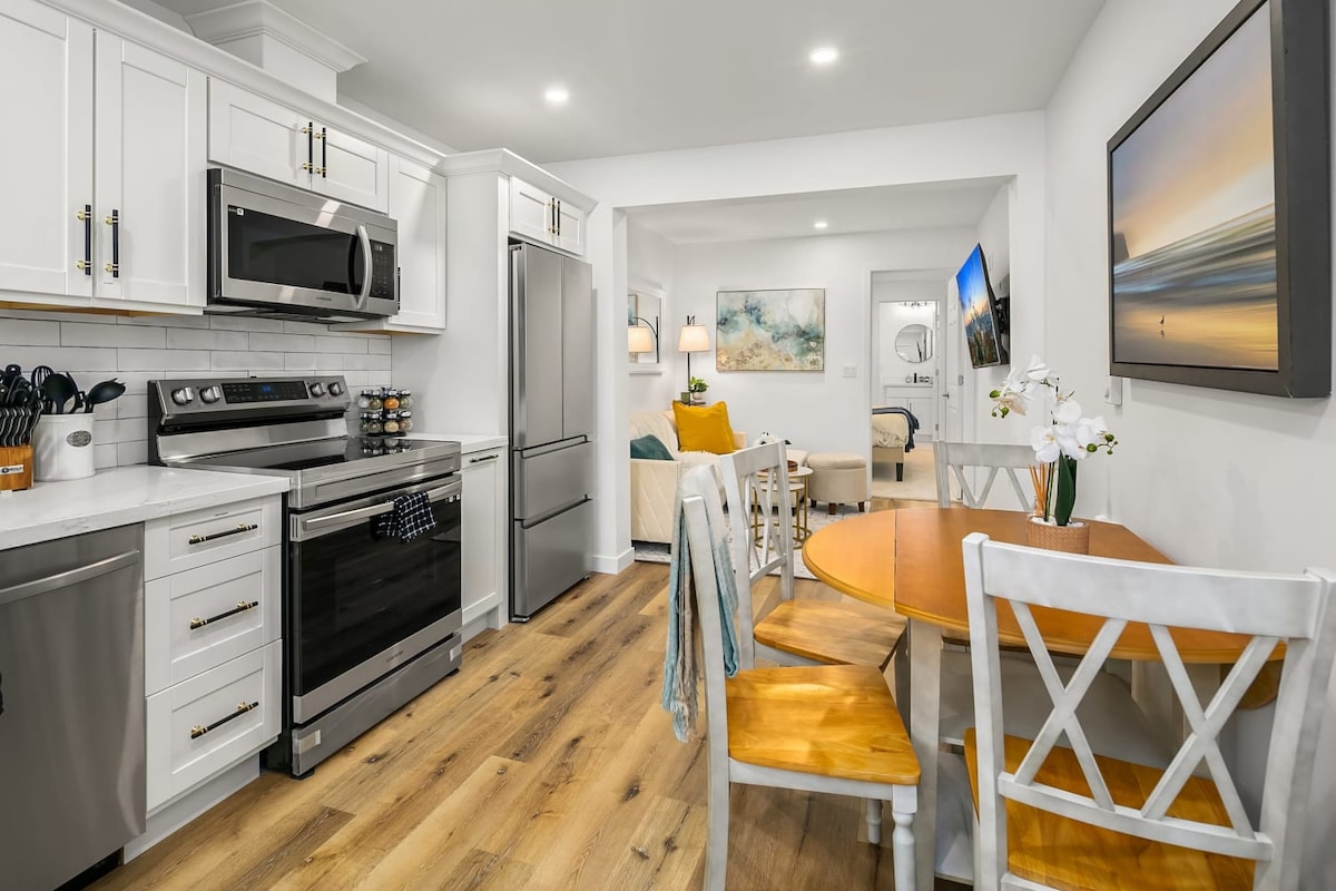 The kitchen features modern stainless-steel appliances, including a microwave and refrigerator. A wooden table with seating for four is positioned nearby, complemented by white cabinetry. The open living area is visible, enhancing the communal space with a comfortable atmosphere.