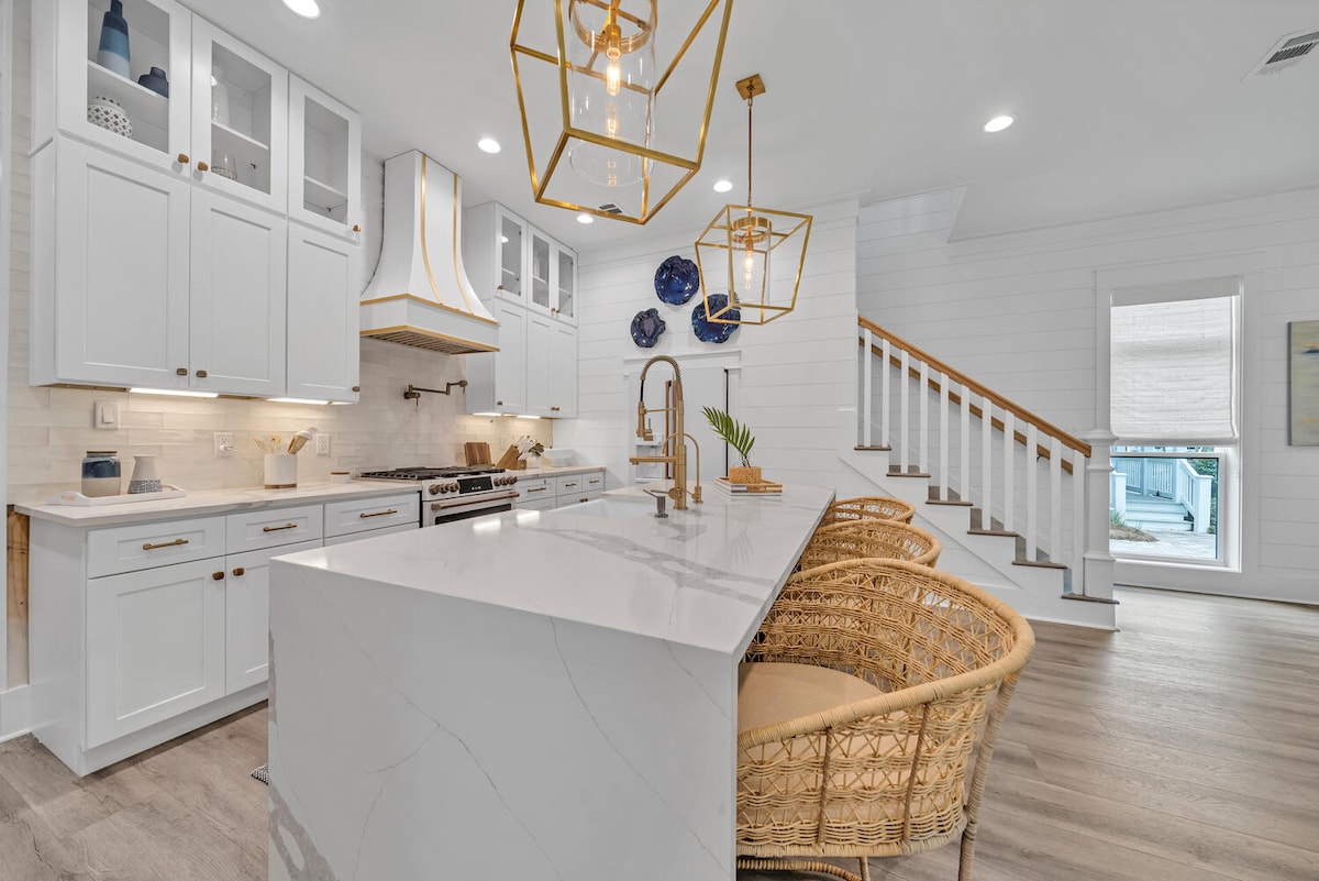 A modern kitchen is highlighted by white cabinetry and a large island with a sleek marble countertop. Rattan bar stools provide seating, while pendant lights with geometric designs hang above. The space features natural light streaming in from nearby windows, enhancing the warm atmosphere.