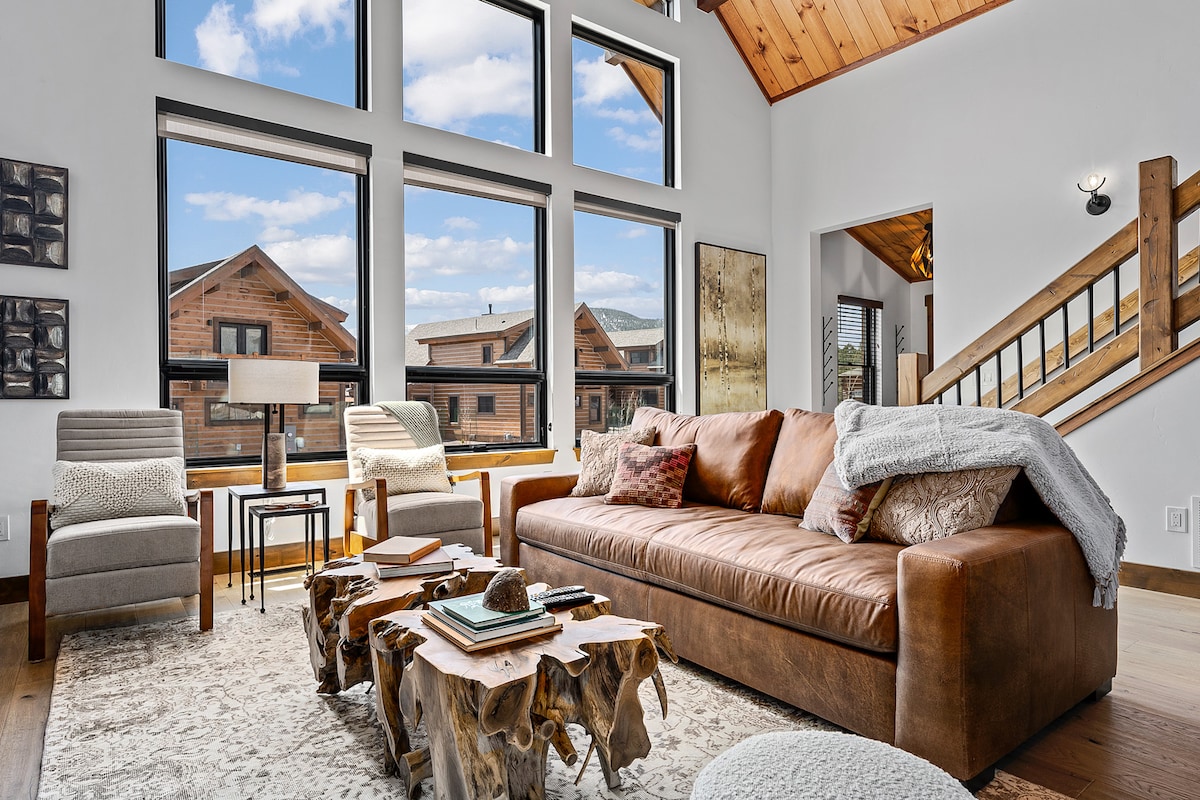 The living area showcases a spacious leather couch complemented by a textured area rug. Two armchairs are positioned near a small side table, while large windows provide ample natural light and views of the surrounding landscape. The ceiling features wooden beams, enhancing the cabin’s rustic charm.