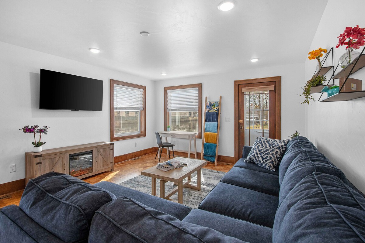 The living room features a large sectional couch facing a flat-screen TV mounted on the wall. A coffee table with magazines rests in front of the couch. Natural light filters through the windows, highlighting the wooden accents and a small dining table in the corner.