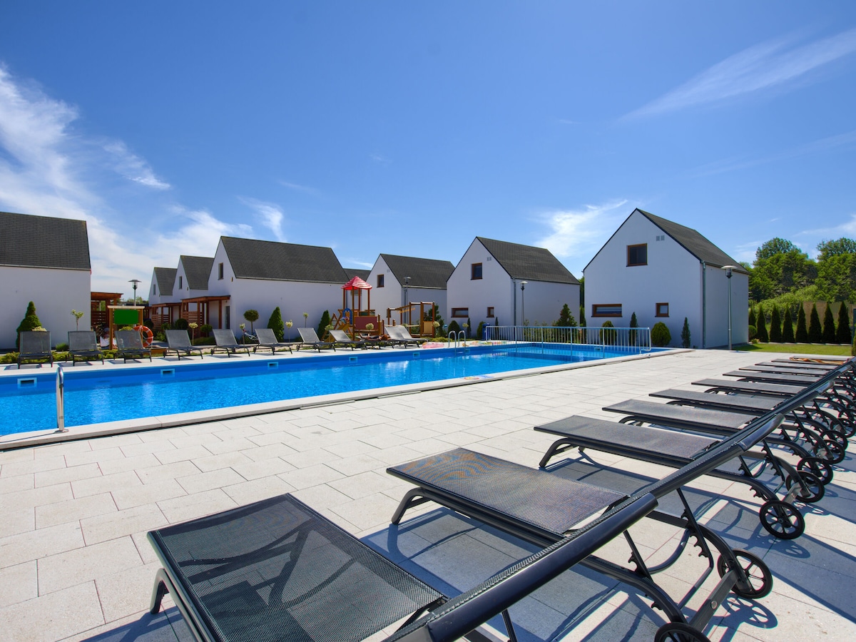 A communal outdoor pool is bordered by spacious sun loungers arranged neatly along the poolside. In the background, modern holiday homes are visible, framed by green landscaping and a clear blue sky.