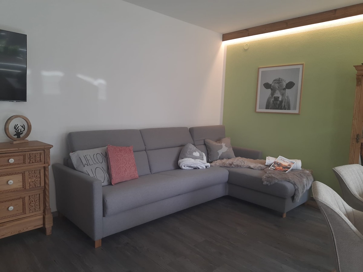 A spacious gray sectional sofa combines comfort with style, accented by decorative pillows and a soft throw. A wooden chest of drawers adds charm to the area, while a framed artwork depicting a cow enhances the relaxing atmosphere. Natural light filters into the room.