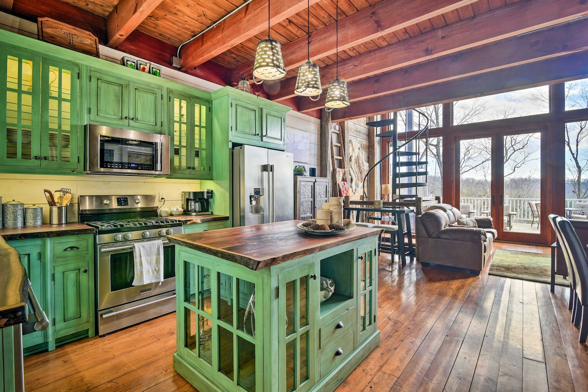 An open kitchen area features green cabinetry with glass panel doors and stainless steel appliances. A wooden island with seating is centered in the space. Natural light enters through large windows, and a spiral staircase adds dimension to the room.