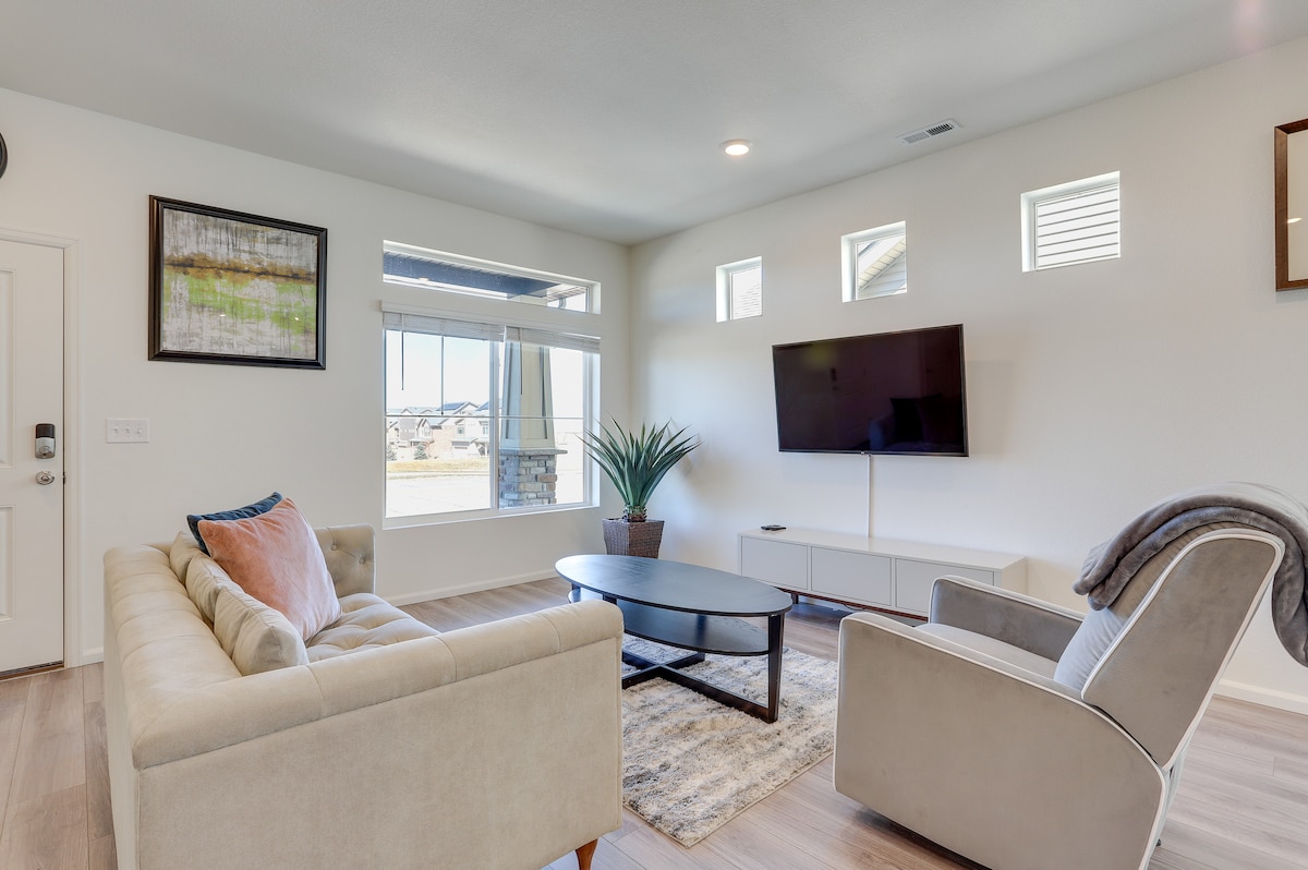 A light-filled living area features a cozy sofa and an armchair, accented by soft throw blankets. A low coffee table sits at the center, complemented by a large television mounted on the wall. Windows above allow natural light to brighten the space.