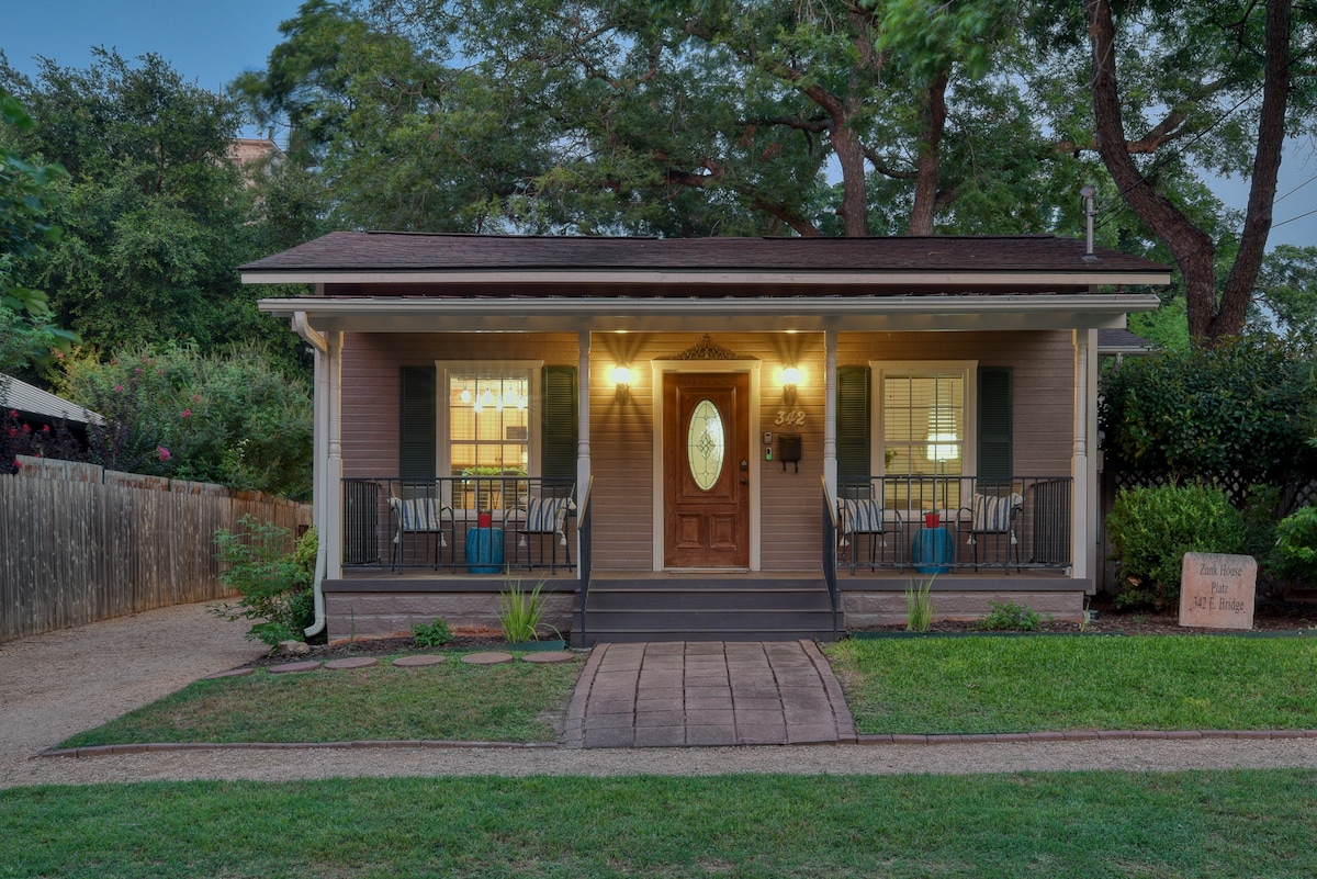A charming cottage exterior is framed by lush greenery and a manicured lawn. Two welcoming porches are adorned with comfortable seating, illuminated by warm exterior lights. The entrance features a decorative door with a circular window, enhancing the inviting appearance of the home.