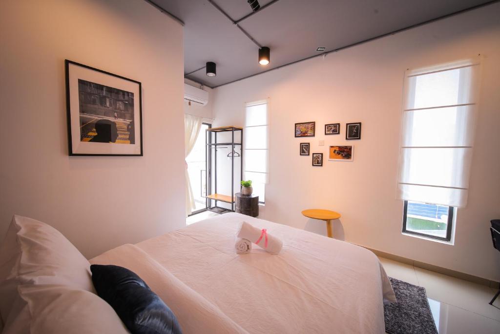 A bright sleeping area is highlighted by a spacious bed with white linens and decorative pillows. Soft light filters through white window coverings, illuminating framed artwork on the walls. A small round table and a plant add functionality and a touch of vibrancy to the space.