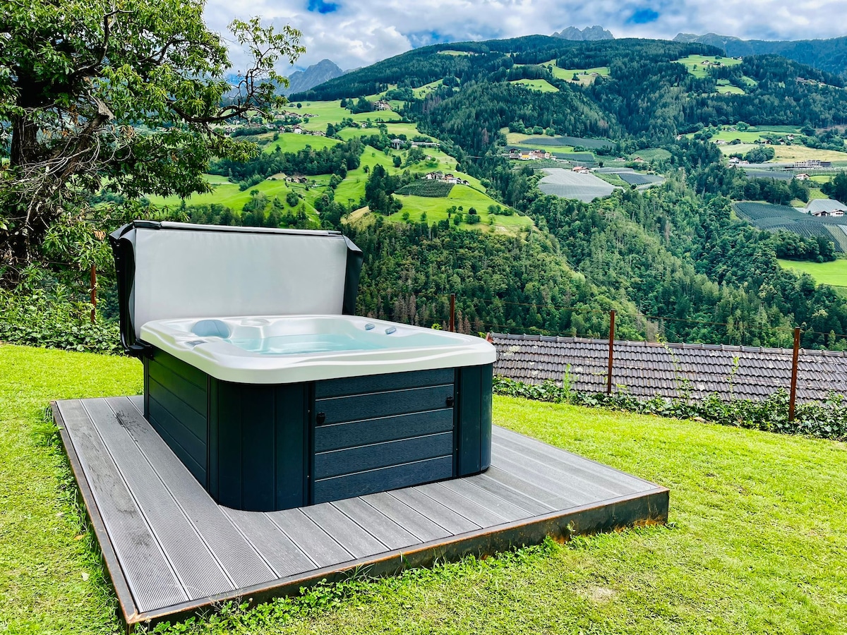 A hot tub is situated on a wooden deck, surrounded by lush greenery and scenic mountain views. The mountains in the background are partially covered by clouds, creating a serene atmosphere for relaxation.