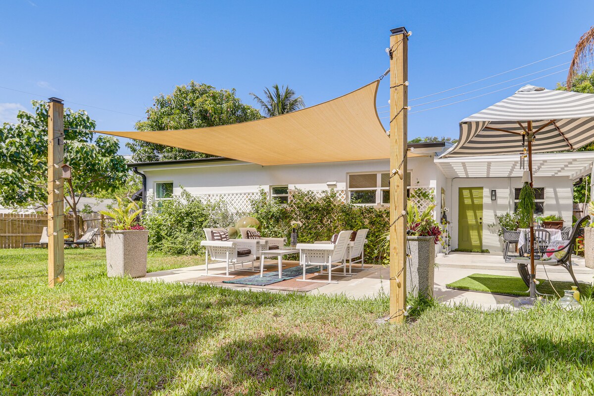 An outdoor patio area is presented, shaded by a large sail. Seating arrangements include comfortable chairs and a table, surrounded by lush green grass and landscaped plant pots, creating a welcoming space for relaxation.