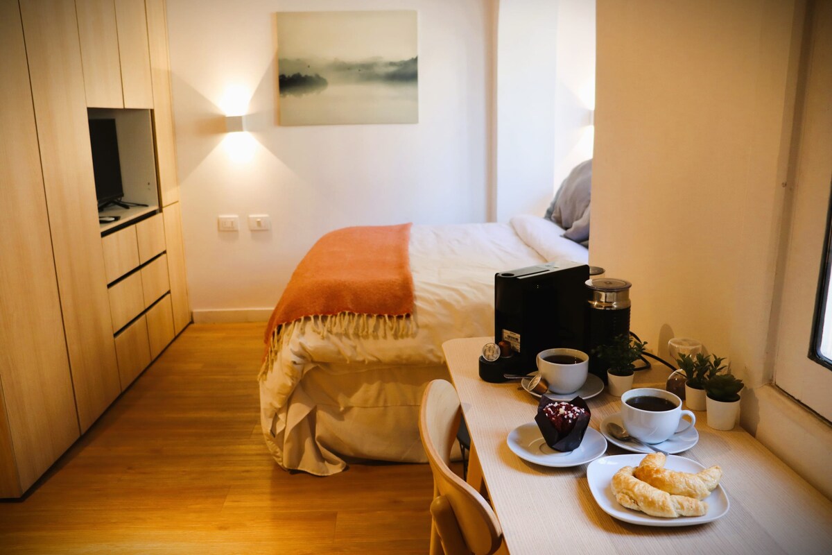 A cozy room is illuminated by soft light, featuring a spacious bed with a warm throw. A small dining table is set with coffee, pastries, and a dessert, alongside a modern coffee maker. A television is mounted on the wall, and plants add a touch of greenery.
