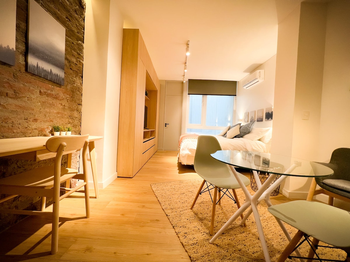 A bright and modern living space features a spacious bed with neutral bedding and natural light filtering through a window. A wooden dining table with two chairs sits beside a glass table with three chairs. A textured wall adds character to the cozy atmosphere.