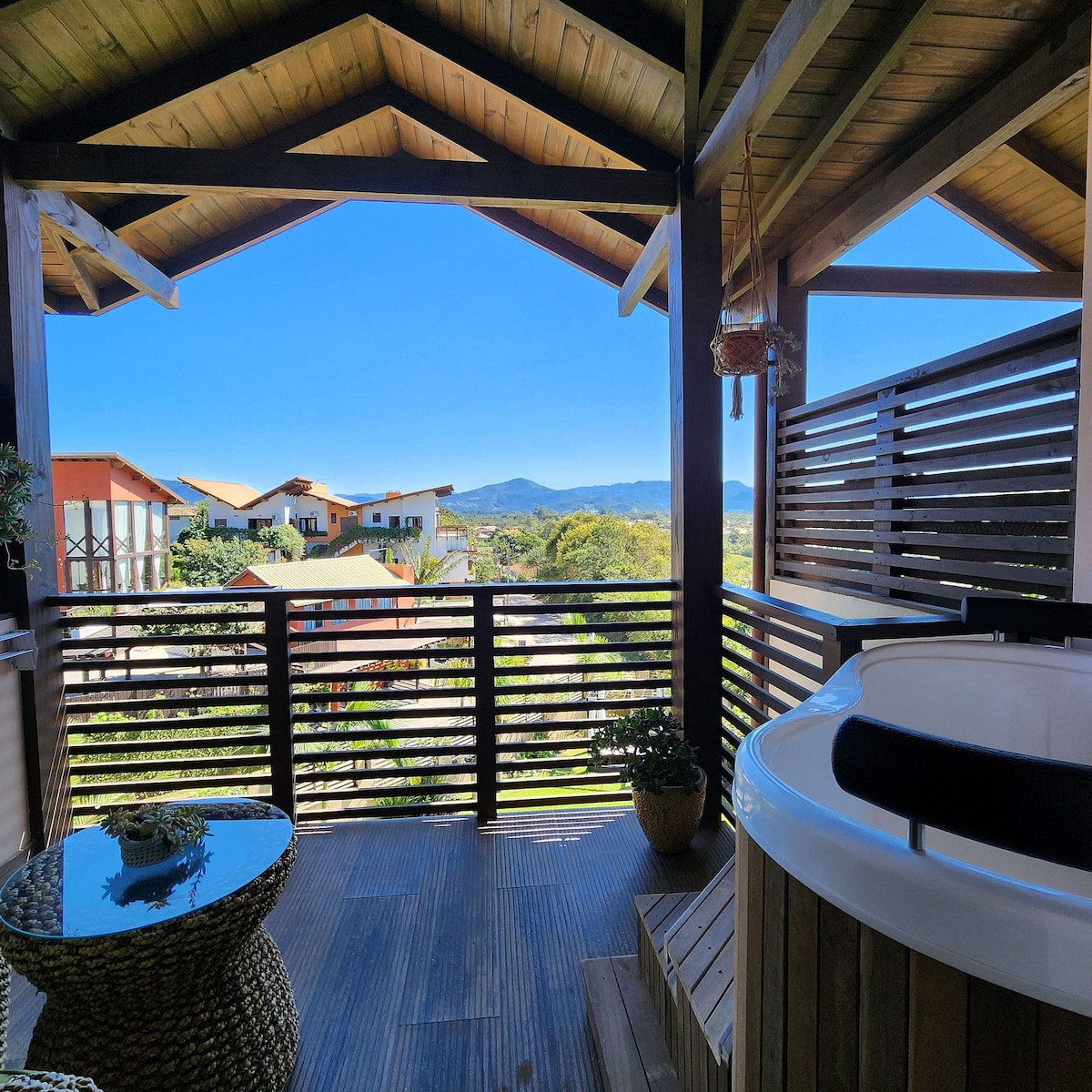 A balcony offers a scenic view of the surrounding landscape, including distant mountains and nearby buildings. A modern hot tub is placed beside a small, woven coffee table, enhancing the serene outdoor space adorned with potted plants.