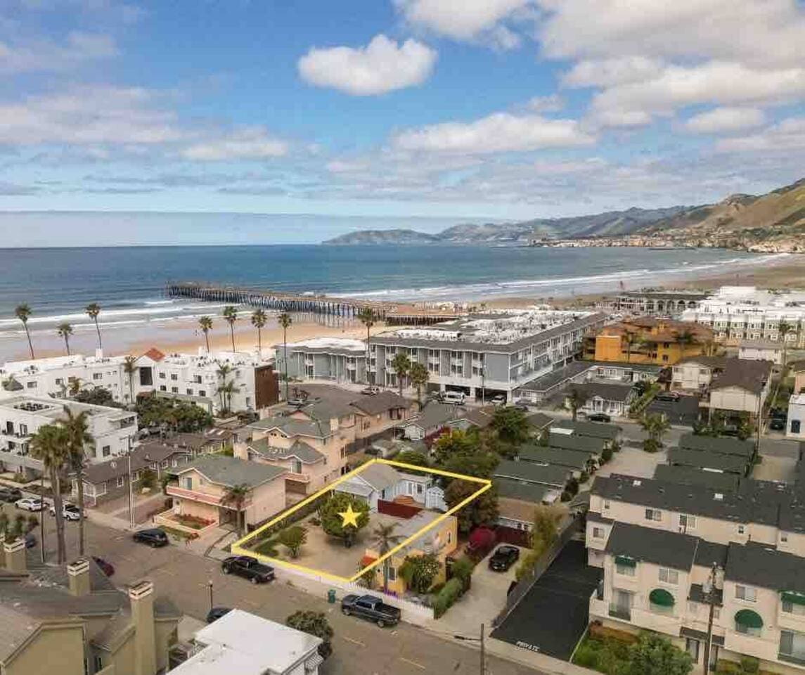 An aerial view captures a coastal neighborhood with charming homes surrounding a fenced property. The ocean glistens nearby, with the pier visible in the distance. Palm trees line the streets, and various structures are positioned to showcase a vibrant beachfront community.