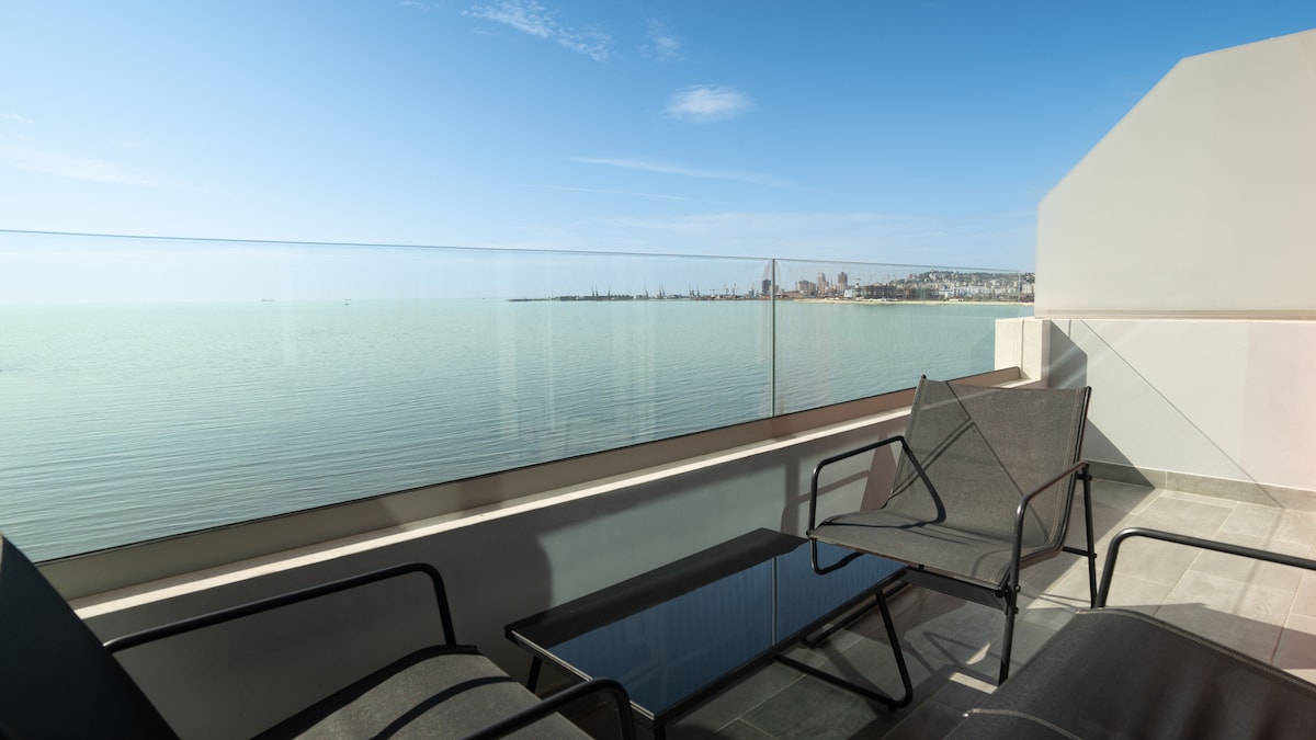 An outdoor terrace features sleek black lounge chairs and a glass table, providing a space to relax. A clear railing offers unobstructed views of the calm sea and distant coastline under a bright blue sky.