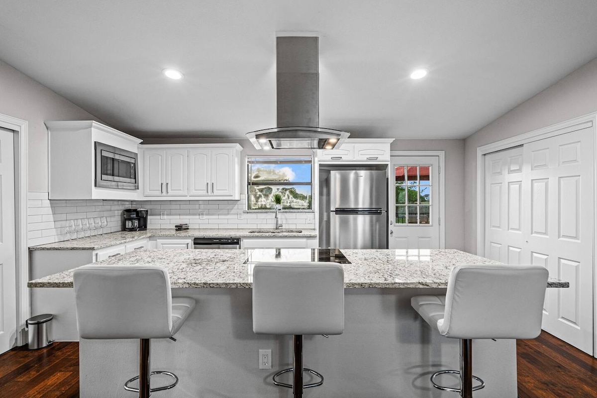 A modern kitchen features a large island with white bar stools and elegant granite countertops. White cabinetry complements stainless steel appliances, including a refrigerator and built-in microwave. A window above the sink offers a view outside, while the open layout enhances spaciousness.