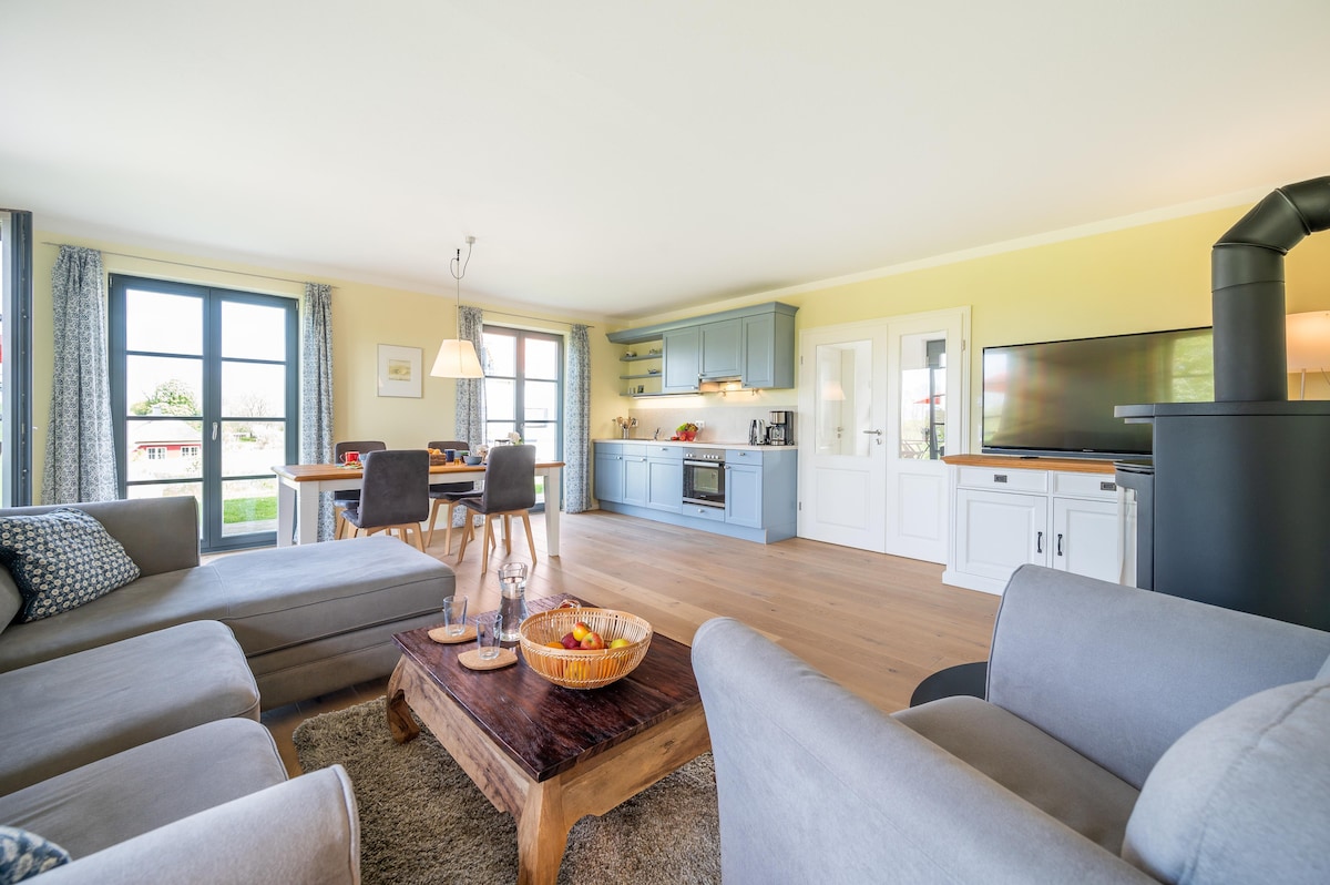 An open living area features a comfortable seating arrangement with gray sofas and a wooden coffee table holding a bowl of fruit. A kitchen area with blue cabinetry is visible, accompanied by a dining table. Large windows allow natural light and provide views of the outside.