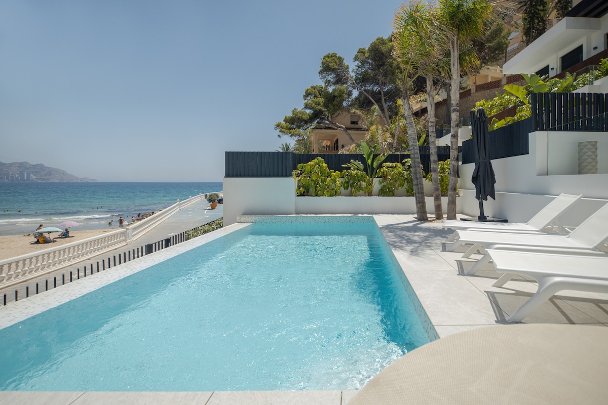 A private pool is visible in the foreground, with clear blue water reflecting the sky. White lounge chairs are arranged around the pool, while lush greenery frames the space. The beach and ocean can be seen in the background, enhancing the coastal setting.