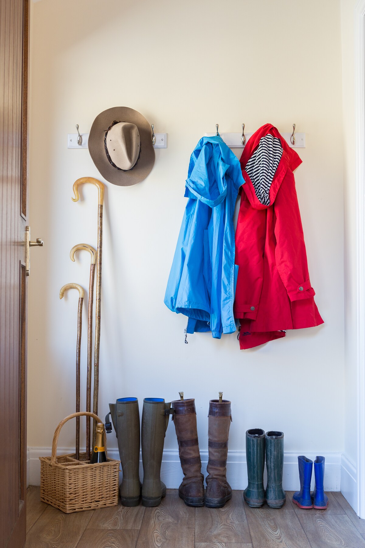A well-organized entryway features hooks holding a blue rain jacket and a red rain jacket, accompanied by a striped hat. A cane stand is arranged alongside a wicker basket. Various pairs of boots, including brown, green, and blue, are neatly lined up on the floor.