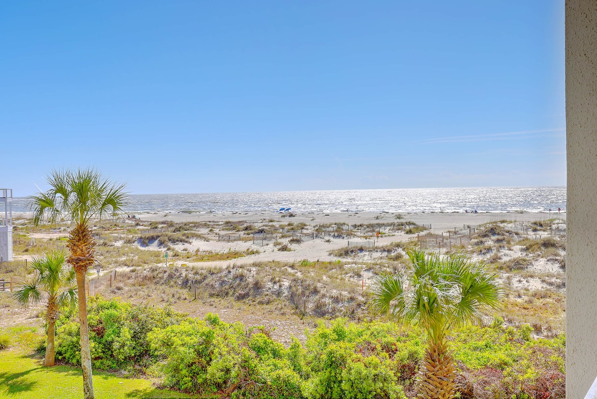 An expansive view of the ocean is visible, featuring soft sandy shores and waves gently rolling in. Lush greenery and palm trees frame the foreground, contributing to a serene beach atmosphere under a bright blue sky.