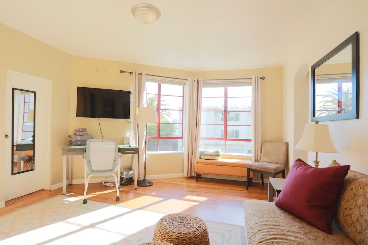 The main living area features a comfortable couch with decorative pillows, a desk with a chair, and a mounted television. Large windows allow natural light to fill the space, complemented by light-colored walls and warm wood flooring.