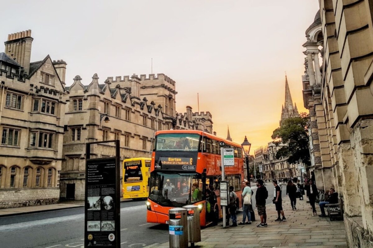 An orange double-decker bus is parked at a bus stop, with passengers boarding and alighting. Historic stone buildings line the street, showcasing intricate architectural details. A warm sunset casts a soft glow over the scene, contributing to a vibrant urban atmosphere.