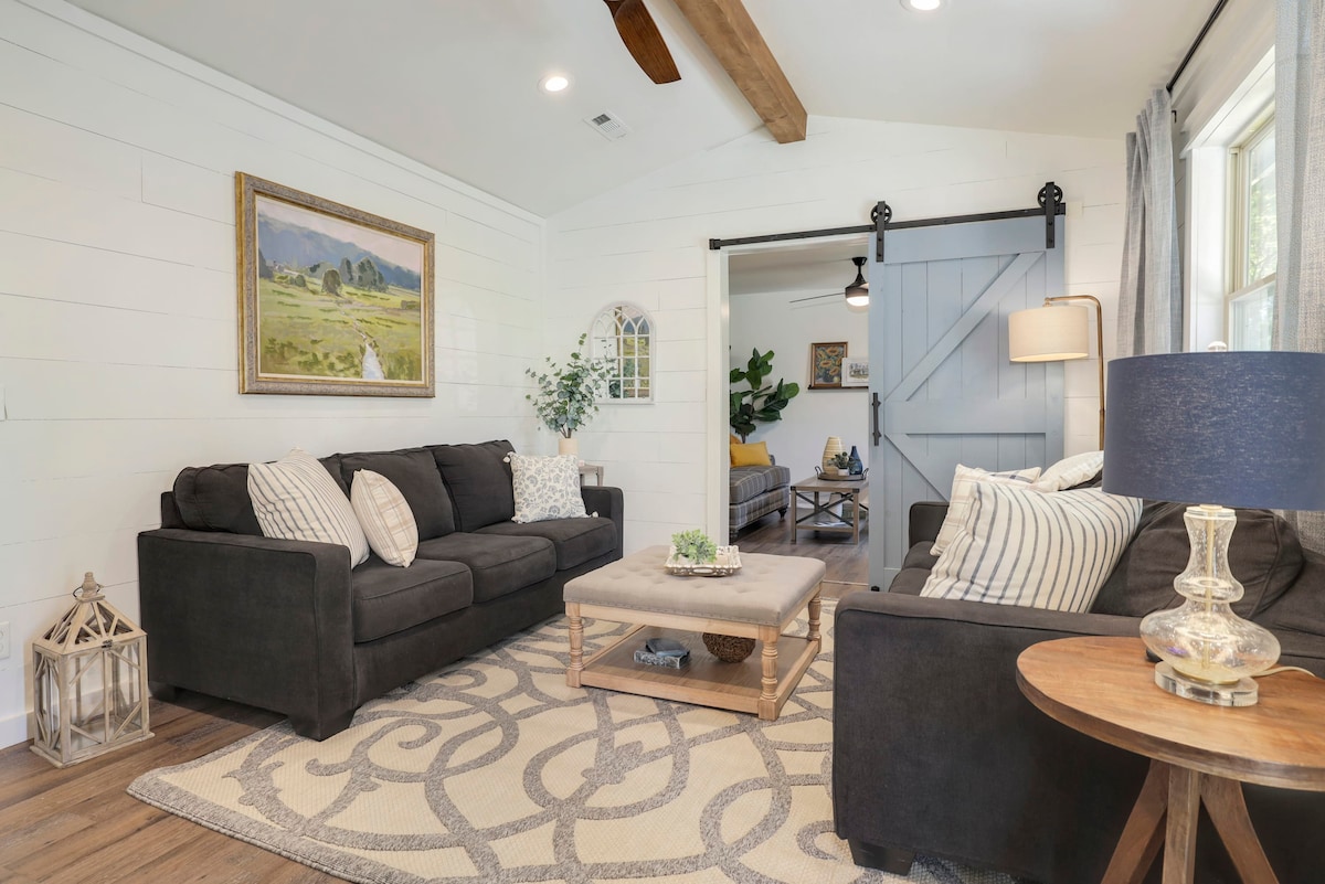 A cozy living room features two dark sofas with decorative pillows facing a central coffee table. A patterned area rug covers the wood floor. Natural light enters through large windows, while a sliding barn door adds a rustic touch. A floor lamp stands beside a side table.