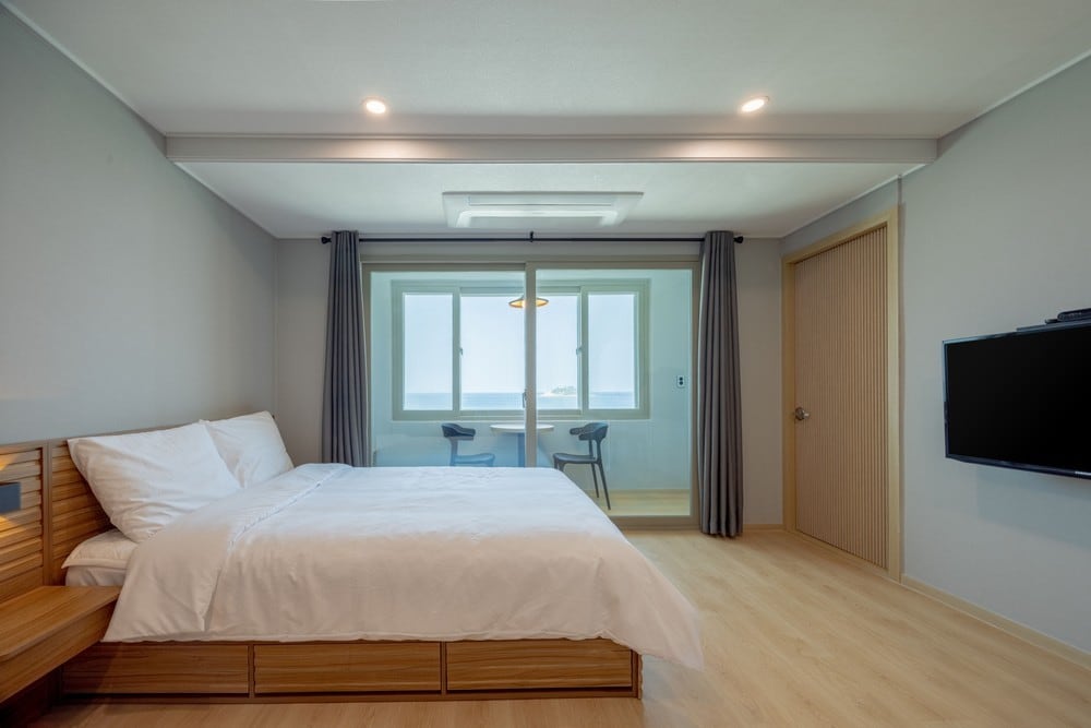 A spacious room contains a large bed with crisp white linens, complemented by wooden accents. Large windows allow natural light to fill the space, offering a view of the ocean. A small seating area with two chairs is positioned near the windows.