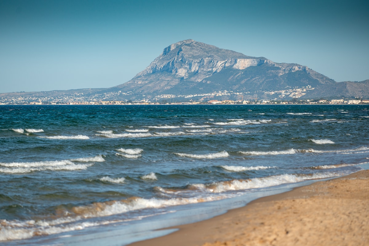 The image captures a serene beach scene with gentle waves lapping at a sandy shore. In the background, a prominent mountain rises against a clear sky, contrasting with the deep blue water. Coastal areas are visible, enhancing the natural scenery.