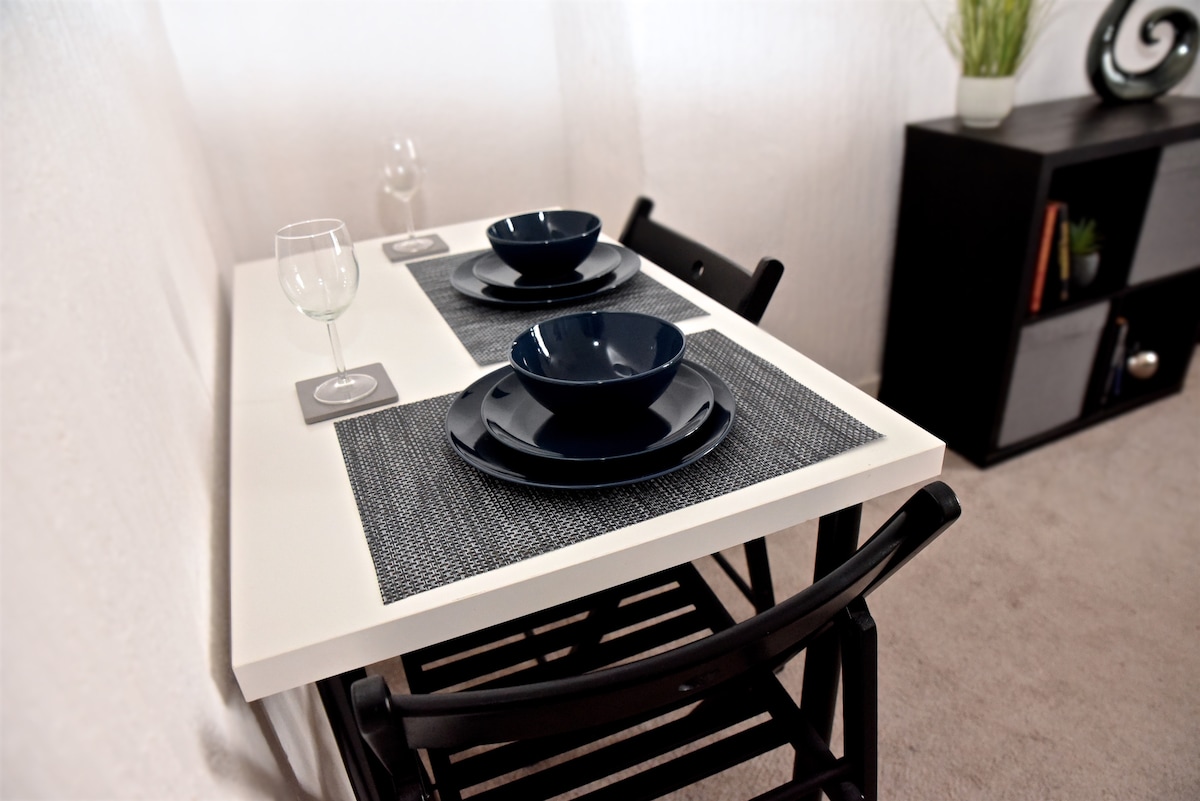 A neatly arranged dining table features two dark plates, two matching bowls, and a wine glass on woven place mats. Black folding chairs are tucked under the table. A stylish shelf with decorative items is visible in the background.