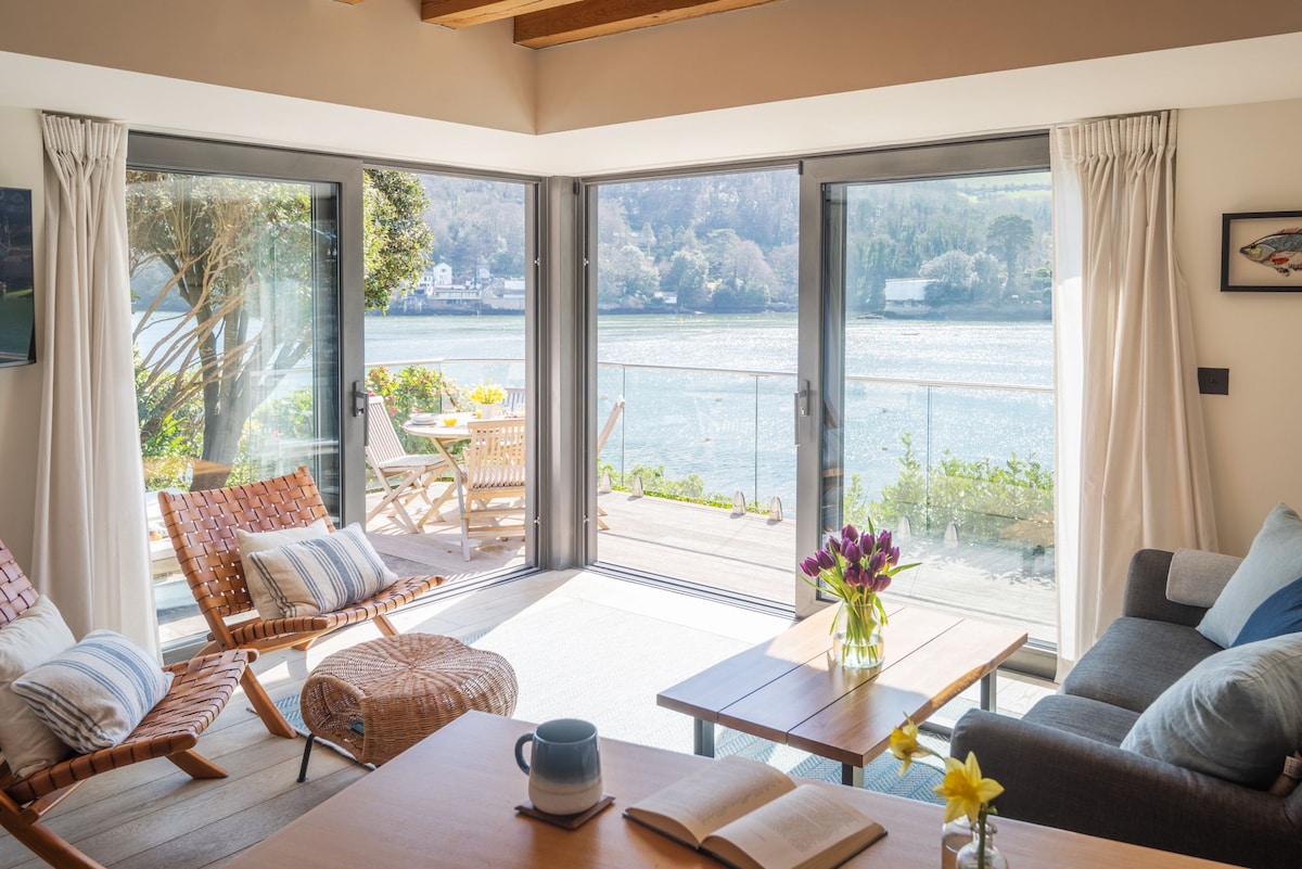 A welcoming living area is filled with natural light, featuring large glass doors that open to stunning views of the River Dart. A wooden table sits in the center, adorned with vibrant flowers. Comfortable seating options include a rattan chair and a plush sofa.