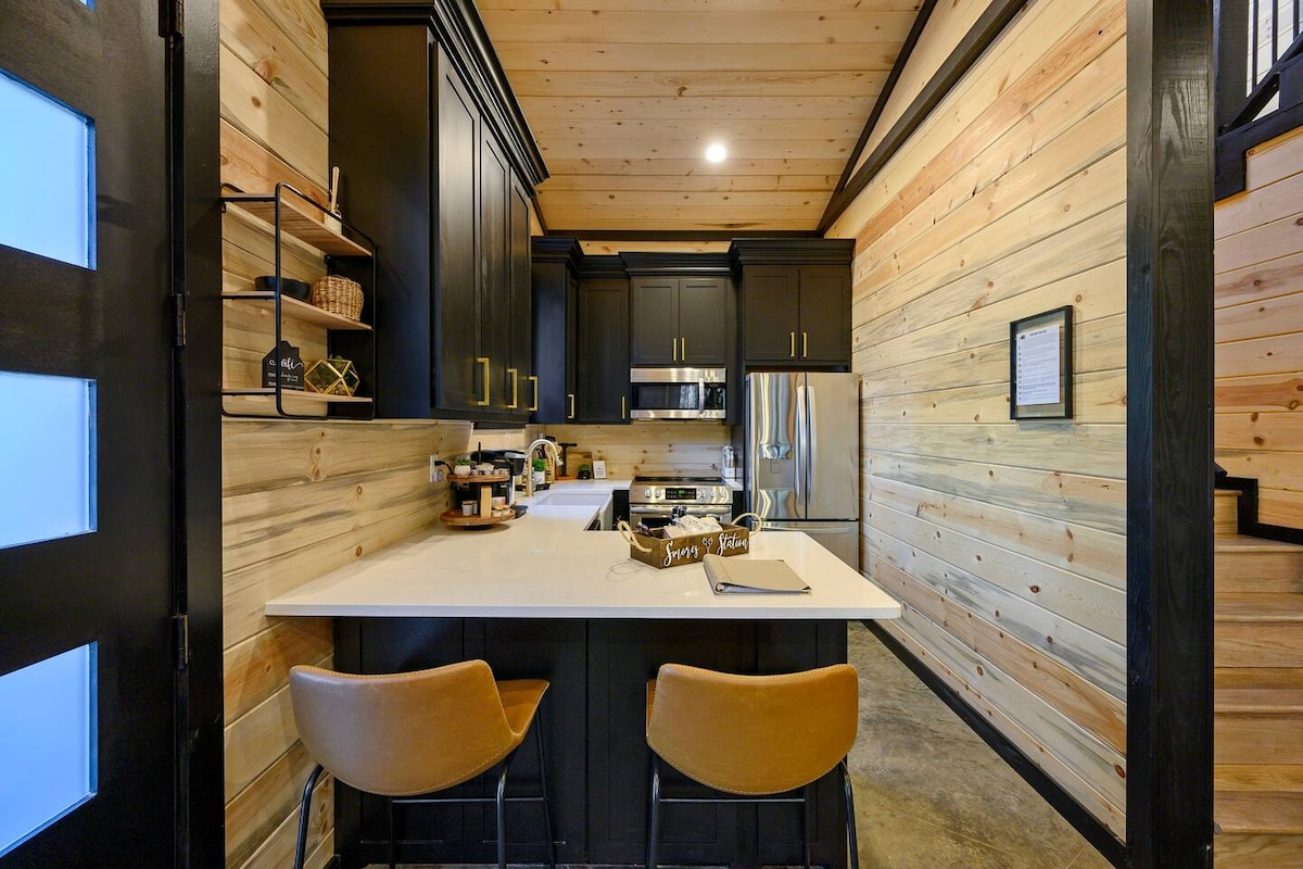 The kitchen showcases modern black cabinetry accented with warm wood paneling. A spacious white island provides seating with two stylish bar stools. Stainless steel appliances reflect light, and countertop essentials are neatly arranged for convenience.