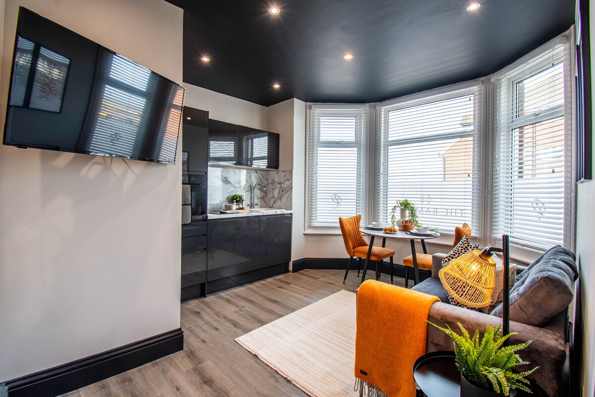 A modern living space features a small dining table with two orange chairs beside a window adorned with white blinds. A plush sofa is complemented by an orange throw. A wall-mounted TV offers entertainment options while light engulfs the room through the large windows.