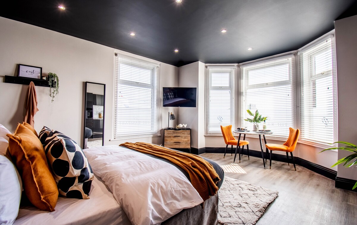 The suite features a spacious sleeping area, with a large bed adorned in neutral linens and contrasting decorative pillows. A wall-mounted TV is positioned above a wooden dresser. A cozy seating area includes two orange chairs paired with a small table, illuminated by natural light from bay windows.