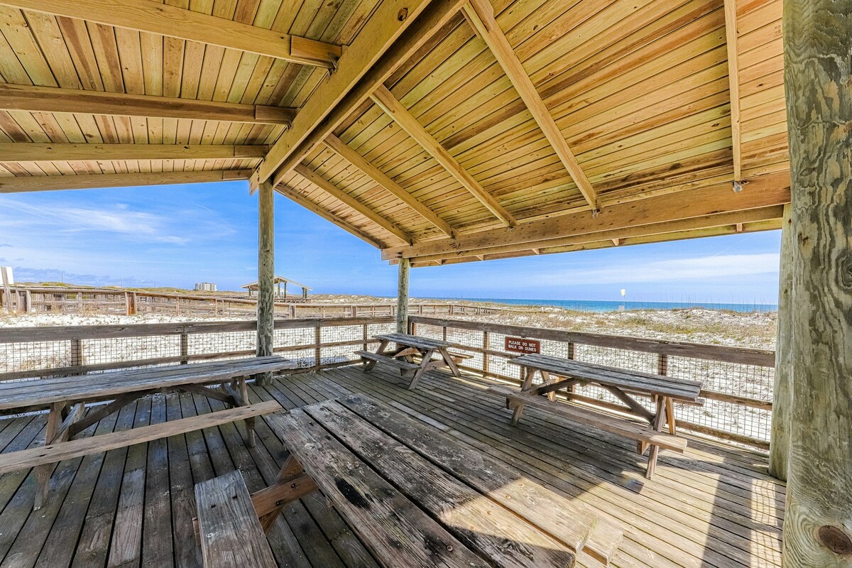 A spacious covered pavilion is shown with wooden picnic tables arranged beneath a high wooden ceiling. The open view showcases a sandy beach and the ocean beyond, bathed in natural light, creating a relaxing setting for gatherings.