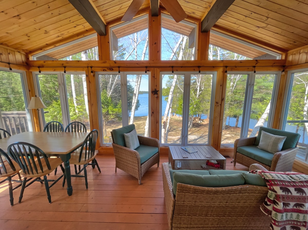An airy living area features expansive windows offering views of the outdoors. A wooden table with six black chairs is positioned next to the windows. A comfortable seating arrangement includes two wicker chairs and a sofa adorned with decorative pillows, enhancing the space's natural light.