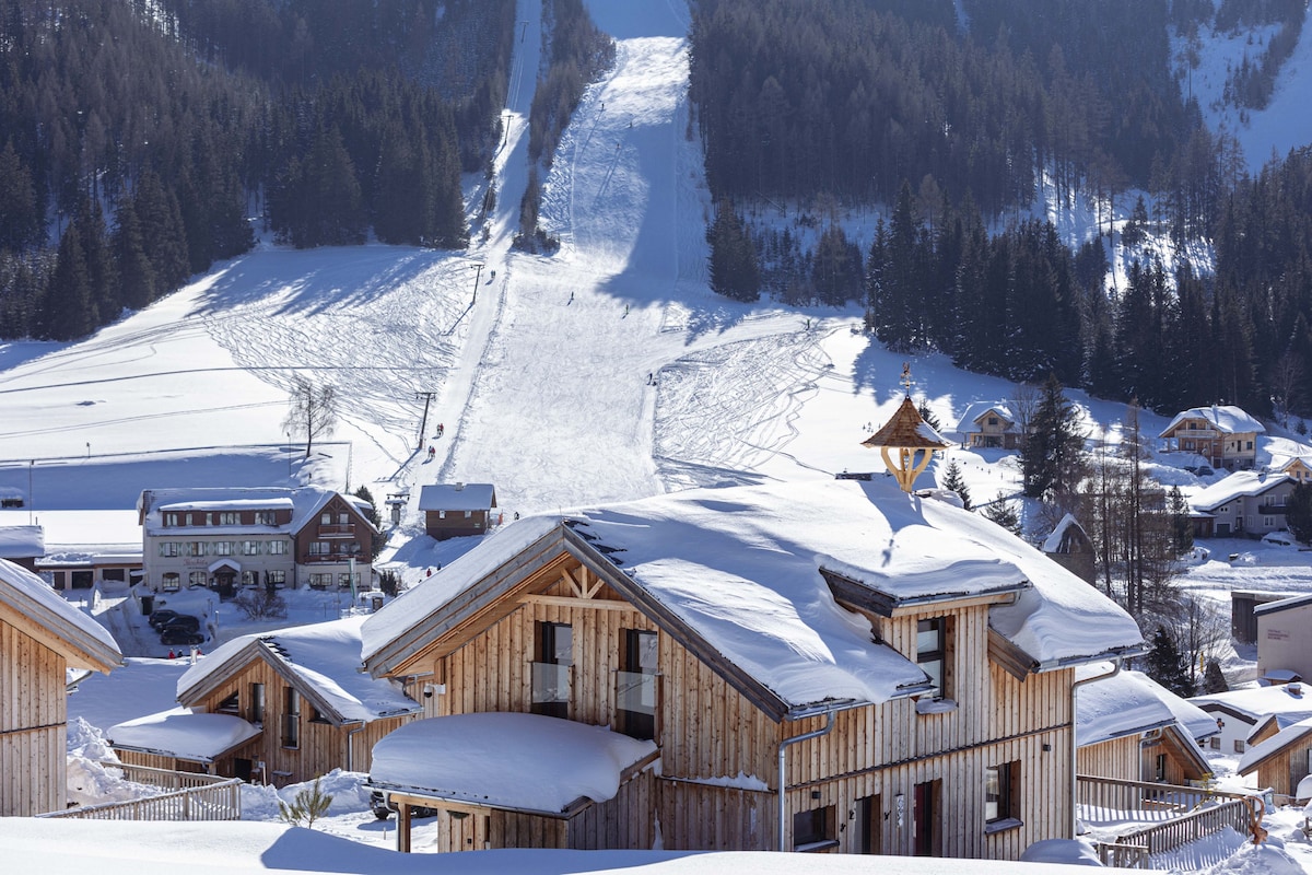 The image captures a scenic view of chalets blanketed in snow, with a ski slope gently descending toward the village in the background. Tall trees frame the landscape, and a clear sky adds to the serene winter atmosphere.