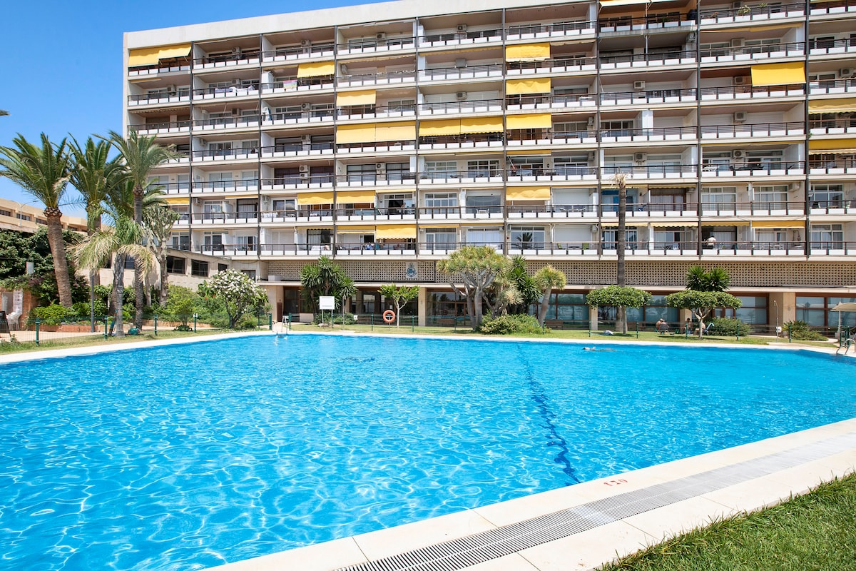A large outdoor swimming pool is surrounded by a well-kept garden area, featuring palm trees and green plants. The multi-story building in the background showcases multiple balconies with yellow awnings and provides access to shared amenities.