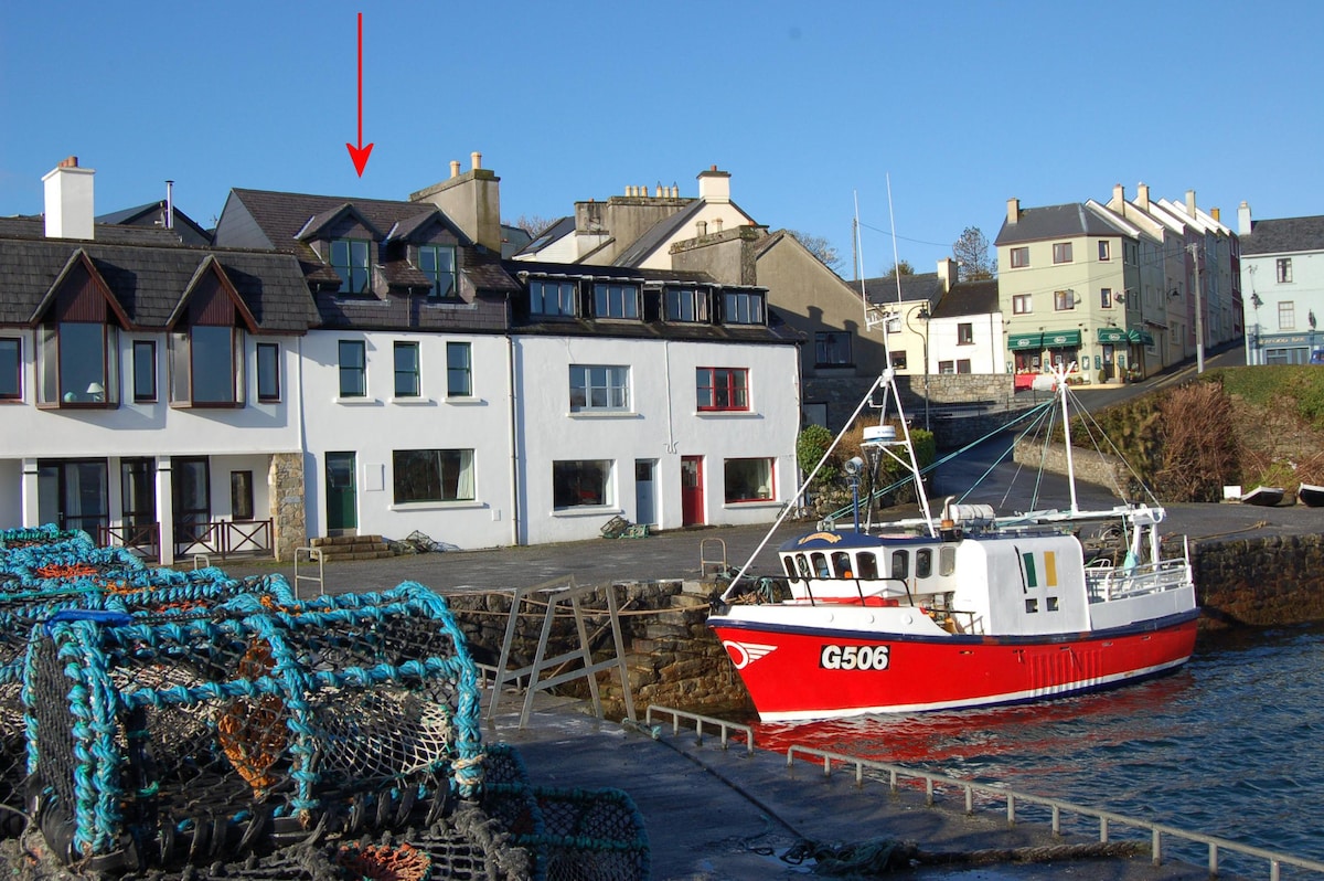The image shows a picturesque coastal scene featuring the Roundstone Quay House and a fishing boat docked by the pier. The house displays a combination of white and dark exterior accents with multiple windows, while the vibrant red boat adds a striking contrast against the water.